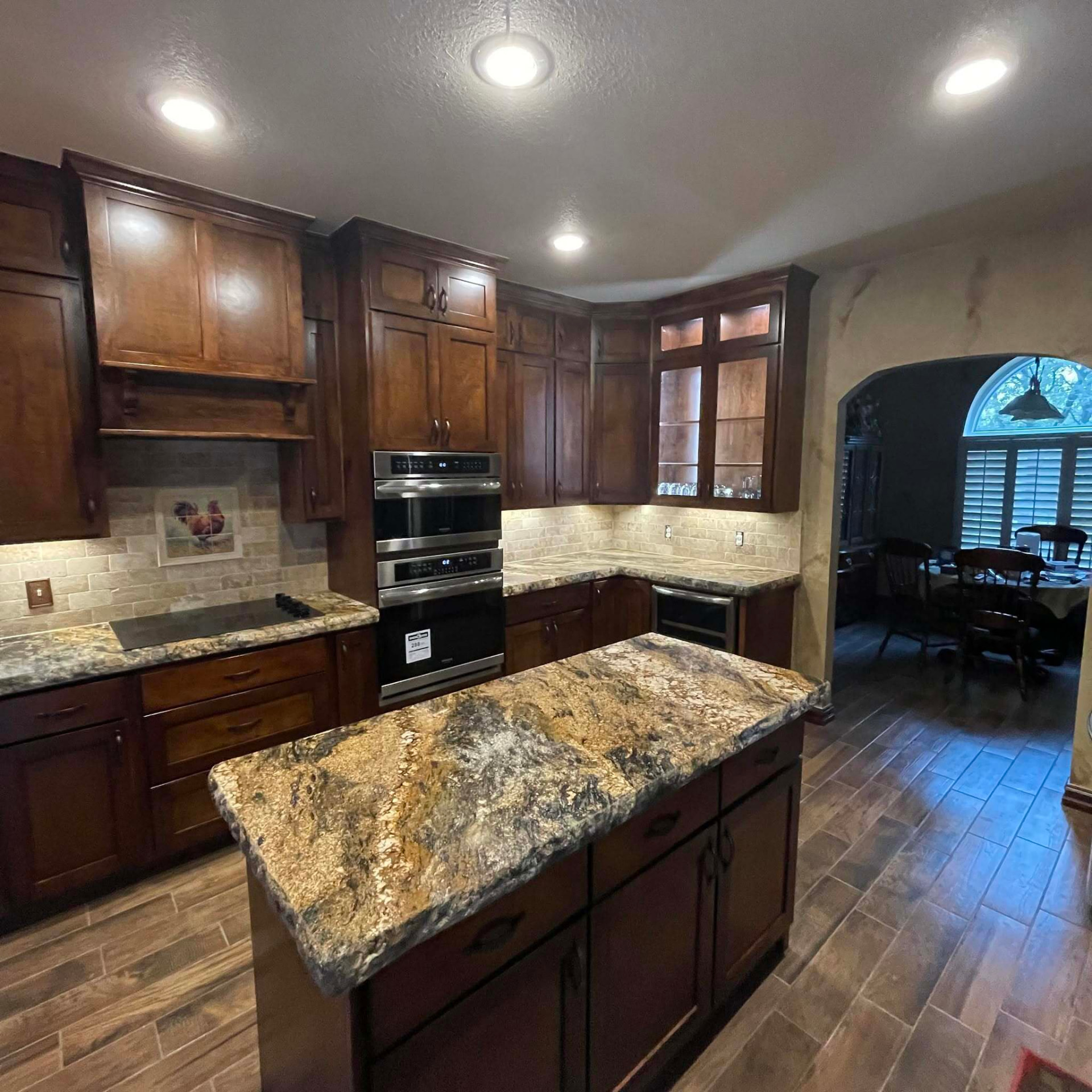 Finished kitchen with dark wood cabinets, granite island, stone backsplash, and wood look tile floor