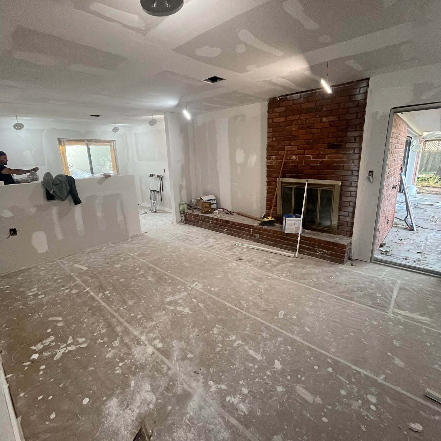 Open living room with fresh drywall hung and taped, brick fireplace exposed, and floors protected